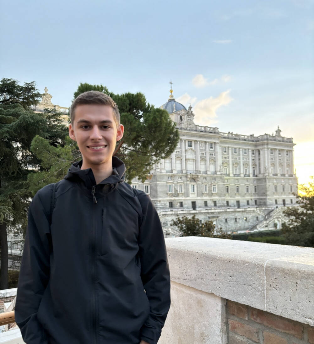 Marcos Sanson, in a black jacket stands outdoors in front of a stone railing, with the grand facade of the Royal Palace of Madrid visible in the background under a partly cloudy sky. Tall trees are also present to the left of the palace.
